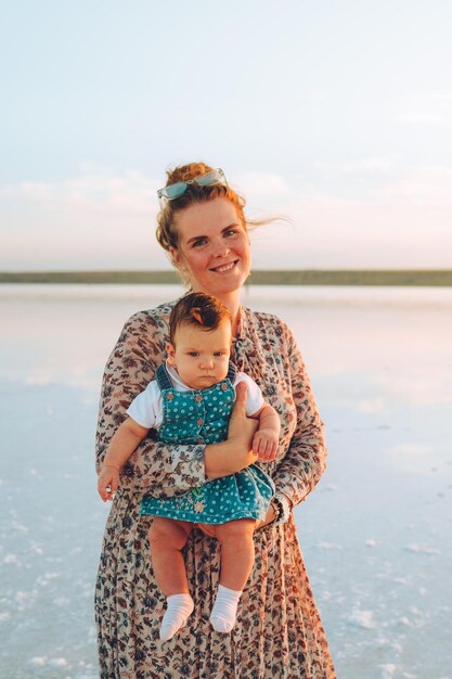 Foto gelukkige glimlachende vrouw met rood haar die haar kind vasthoudt en langs de zee loopt