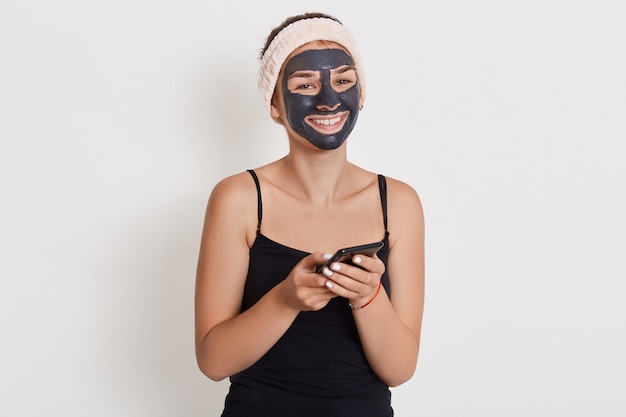 Gelukkige glimlachende vrouw met zwart kosmetisch masker met gelukkige blik, houdend telefoon, controlerend sociaal netwerk terwijl het doen van kosmetische procedures.