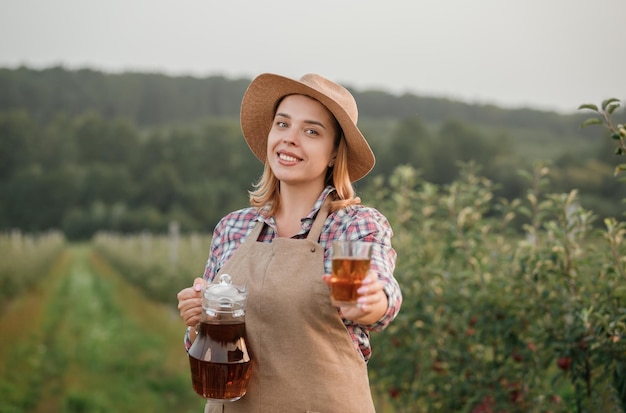Gelukkige glimlachende vrouwelijke landbouwer die smakelijk appelsap in glas giet dat in de boomgaardtuin staat tijdens de herfstoogst Oogsttijd
