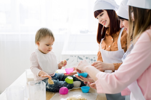 Gelukkige glimlachende vrouwen die samen met weinig keuken van het babymeisje thuis bakken, Moedersdag, Familieconcept. Vrouwen stoppen deeg in gekleurde siliconen bakvormpjes voor muffins