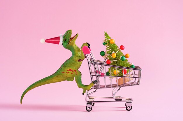 Foto gelukkige groene speelgoeddinosaurus die een winkelwagentje draagt met een kerstboom op een pastelroze achtergrond minimale trendy kunstwagentje voor de wintervakantie