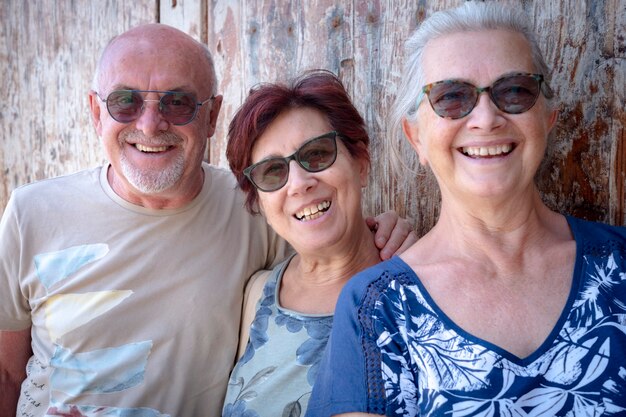 Gelukkige groep van drie vrienden die genieten van een buitenexcursie die op een zonnige dag tegen een houten deur staat - actieve en zorgeloze ouderen die met pensioen zijn en samen plezier hebben