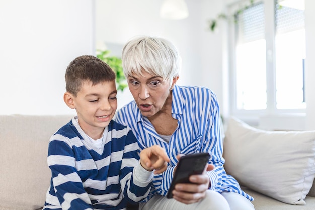 Gelukkige grootmoeder en kleinzoon die samen telefoon gebruiken, thuis op een gezellige bank zitten, door apps voor mobiele apparaten bladeren, oma met kleinkind een spel spelen, naar het scherm kijken, plezier maken