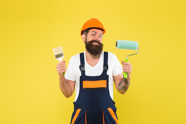 Gelukkige hipster in veiligheidshelm en werkuniform houden verfroller en penseel vast voor decoreren en schilderen in aanbouw gele achtergrond, schilder.