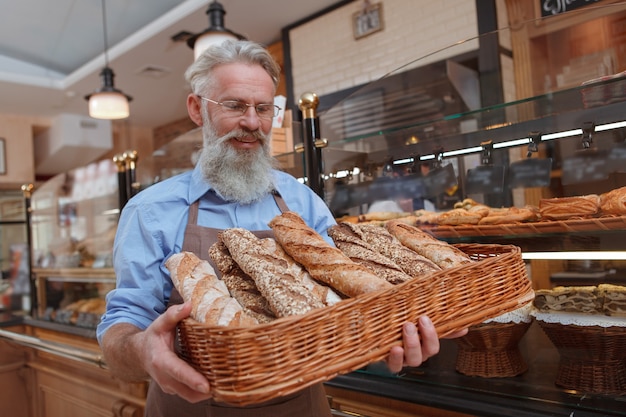 Gelukkige hogere bakker die vreugdevol glimlacht, die vers brood in een mand bekijkt die hij bij zich heeft