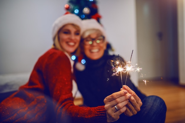 Gelukkige hogere vrouw en haar dochter die nieuw jaar vieren. beiden met kerstmutsen op het hoofd en met sterretjes. op de achtergrond is de kerstboom. familiewaarden concept. selectieve aandacht voor handen.