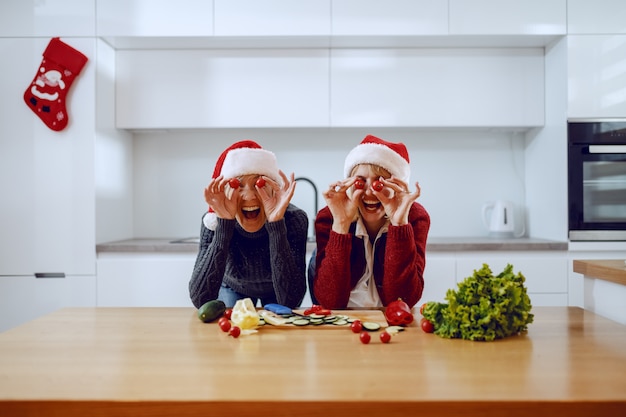 Gelukkige hogere vrouw en haar dochter die op keukenteller leunen en plakken van kersentomaat op ogen houden. Beiden met kerstmutsen op het hoofd. Op het aanrecht staan groenten.