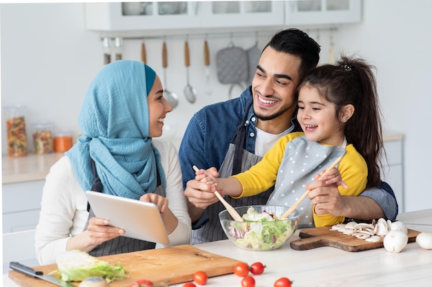 Gelukkige islamitische familie van drie eten recept op digitale Tablet controleren tijdens het koken in de keuken samen, vrolijke moslim moeder, vader en dochtertje genieten van de voorbereiding van de lunch thuis, close-up