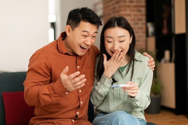 Gelukkige japanse man van middelbare leeftijd en zijn jonge vrouw verheugen zich over een positieve zwangerschapstest in het interieur van de woonkamer