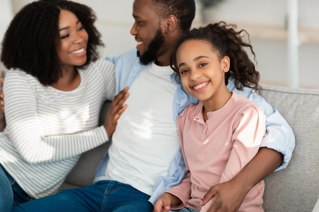 Gelukkige jeugd en liefdevol familieconcept. Portret van lachende zwarte meid zittend met haar knuffelende ouders op de bank in de woonkamer, tijd samen doorbrengen, poseren camera kijken, selectieve focus