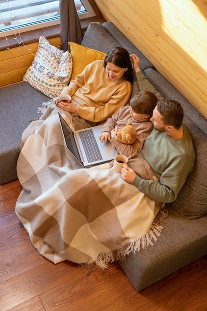 Gelukkige jonge aanhankelijke familie in truien ontspannen op de bank onder geruite plaid in het landhuis en online film kijken