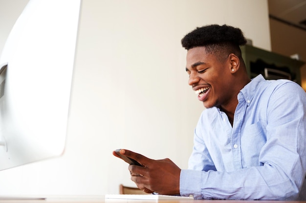 Gelukkige jonge Afrikaans-Amerikaanse man zit met mobiele telefoon bij de computer
