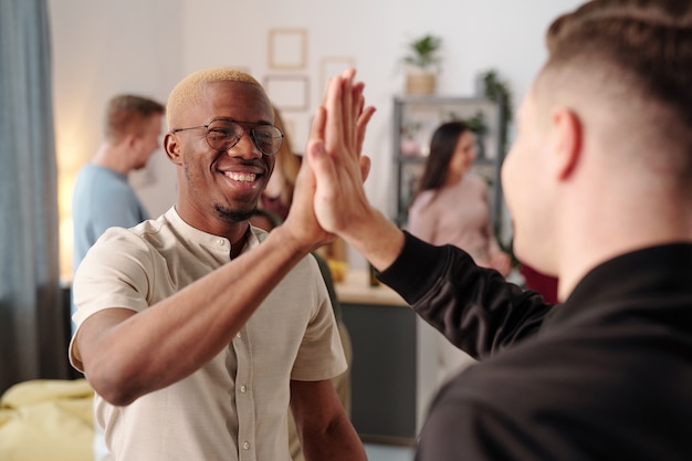 Gelukkige jonge Afrikaanse man in vrijetijdskleding en bril die high five geeft aan zijn blanke vriend op een feestje terwijl hij naar hem kijkt met een brede glimlach