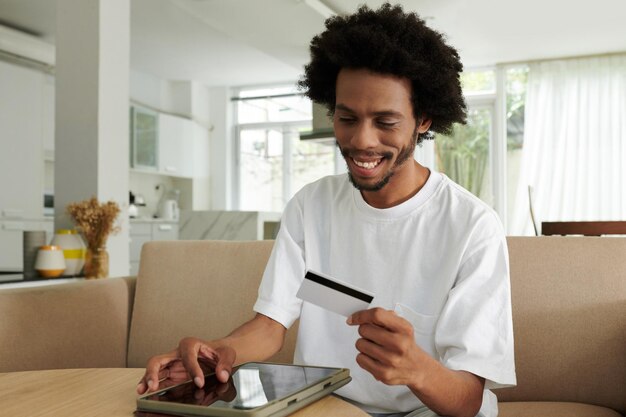 Gelukkige jonge Afro-Amerikaanse man kijkt naar zijn creditcard in zijn hand terwijl hij thuis op de bank zit en het nummer op de website invoert