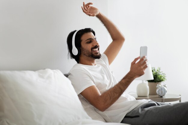 Gelukkige jonge arabische man in witte t-shirt en draadloze koptelefoon met smartphone zit op bed geniet