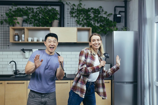 Gelukkige jonge Aziatische man en vrouw die samen zingen en dansen in de keuken, samen plezier hebben in een verliefde familie