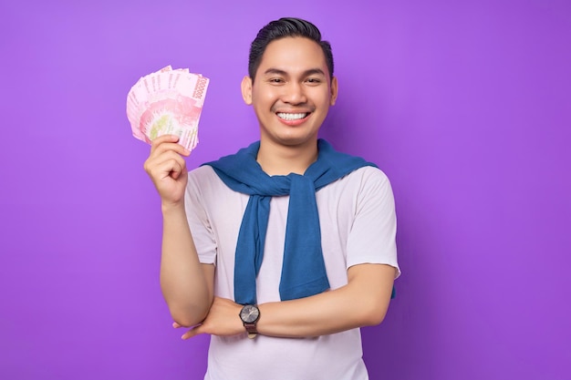 Gelukkige jonge Aziatische man met witte t-shirt casual kleding die geld bankbiljetten aanhoudt en glimlacht naar camera geïsoleerd op paarse achtergrond Winst en rijkdom concept