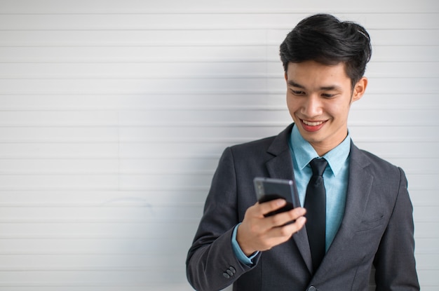 Gelukkige jonge Aziatische manager in pak die lacht en mobiele telefoon doorbladert terwijl hij op een grijze muur staat