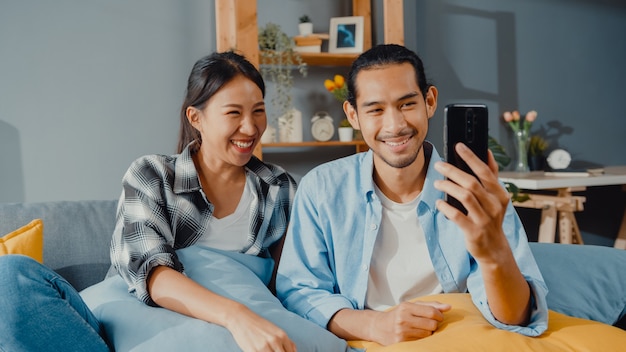 Gelukkige jonge Aziatische paar man en vrouw zitten op de bank gebruik smartphone face-time videogesprek met vrienden en familie