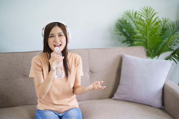 Gelukkige jonge Aziatische vrouw die alleen zingt met een fles als een microfoon en een hoofdtelefoon draagt, houdt van muziek, gelukkig ontspannen in de woonkamer
