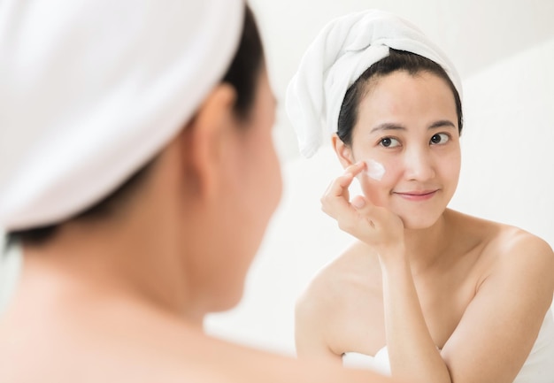 Gelukkige jonge Aziatische vrouw die gezichtslotions aanbrengt terwijl ze een handdoek draagt en haar gezicht in de badkamer aanraakt
