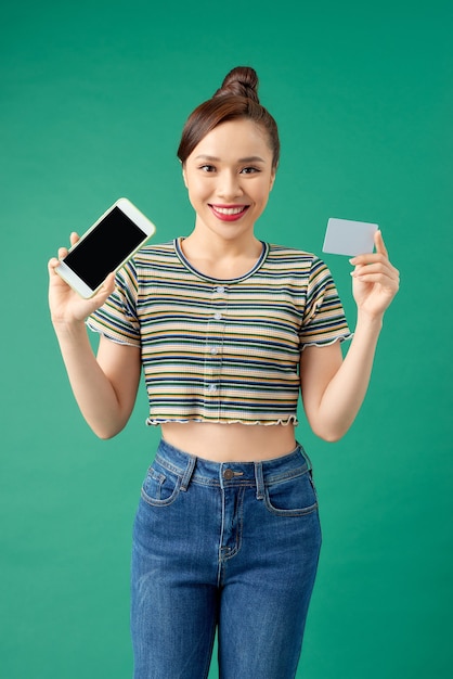 Gelukkige jonge Aziatische vrouw die lege mobiele telefoon en creditcard toont die over groen wordt geïsoleerd.