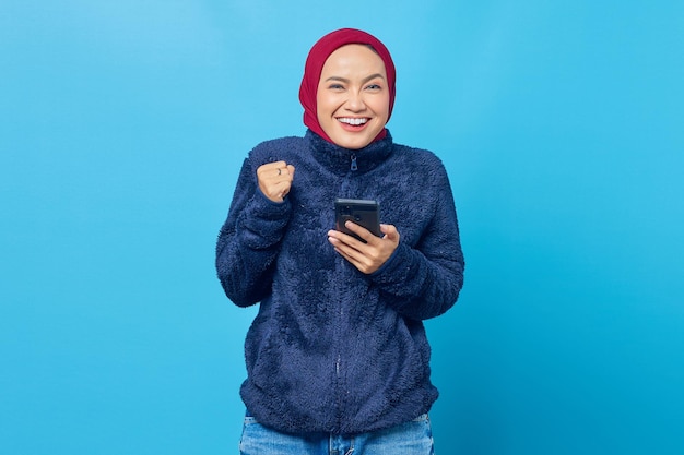 Gelukkige jonge Aziatische vrouw die mobiele telefoon met succesvol handgebaar op blauwe achtergrond gebruikt