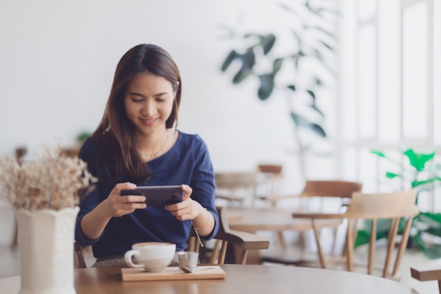 Gelukkige jonge aziatische vrouw die smartphone met het glimlachen gezicht in de winkel van de koffiekoffie gebruiken