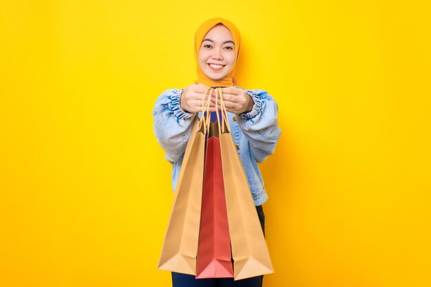 Gelukkige jonge Aziatische vrouw in jeans jasje met shopper tassen en glimlachend naar de camera geïsoleerd over gele achtergrond