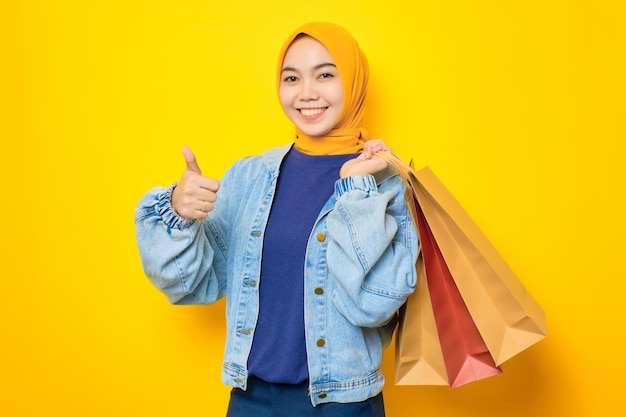 Gelukkige jonge Aziatische vrouw in spijkerjasje met boodschappentassen en gebarende duimen omhoog die geweldige verkoop aanbeveelt, geïsoleerd op gele achtergrond
