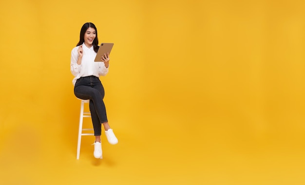 Gelukkige jonge Aziatische vrouw met een glimlach die een tablet gebruikt terwijl ze op een witte stoel zit en kijkt