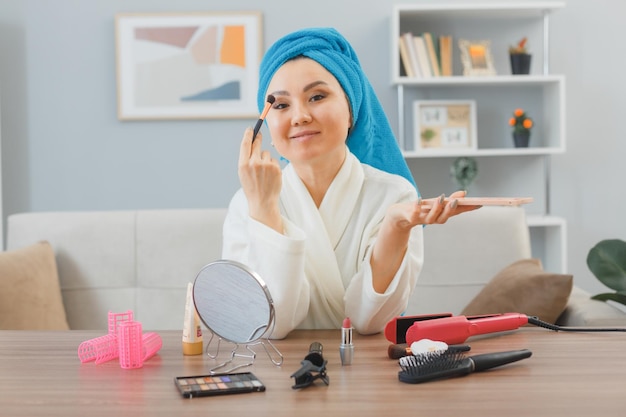 gelukkige jonge aziatische vrouw met een handdoek op haar hoofd zittend aan de kaptafel thuis interieur aanbrengen van oogschaduw in spiegel kijken doen ochtend make-up routine schoonheid en gezichtscosmetica concept