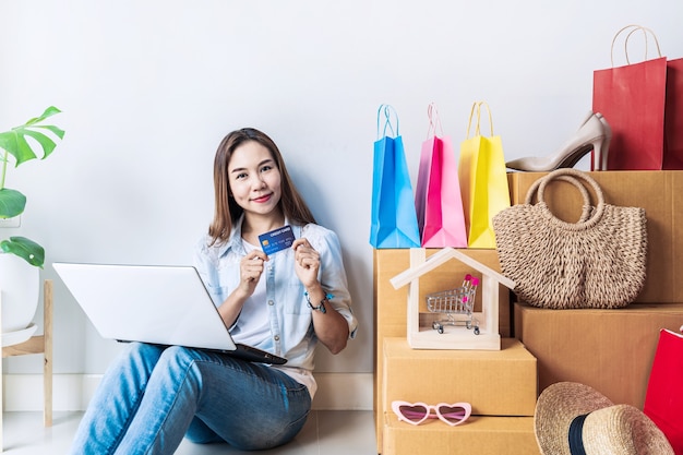 Gelukkige jonge aziatische vrouw met kleurrijke boodschappentas, modeartikelen en stapel kartonnen dozen thuis, met creditcard voor online winkelconcept