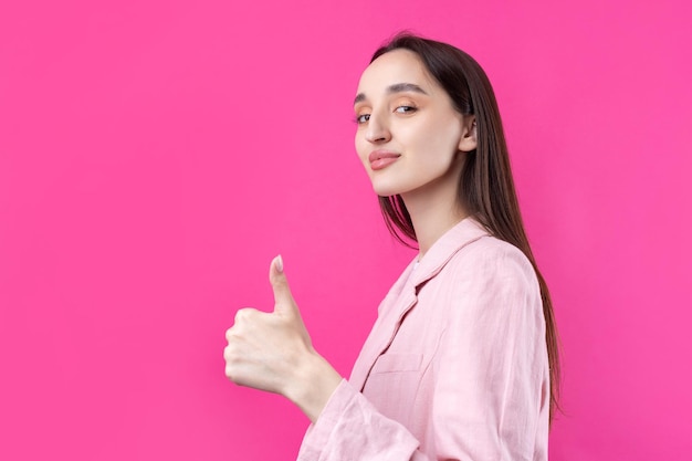 Gelukkige jonge blanke vrouw in een roze jasje die duim omhoog tekent en glimlacht Goed werk en respect