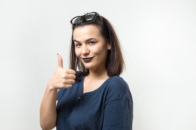 Gelukkige jonge blanke vrouw met een bril en een blauwe t-shirt die duim omhoog tekent en glimlacht