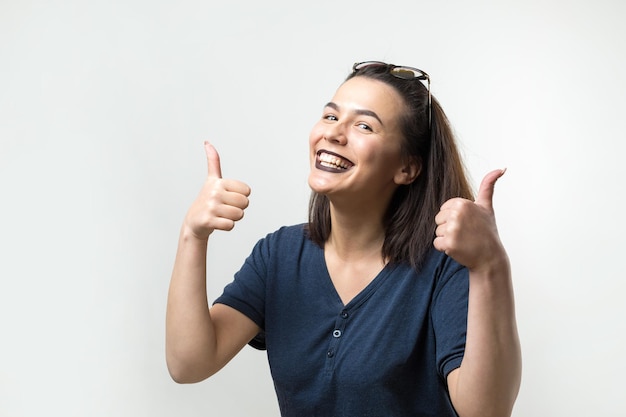 Gelukkige jonge blanke vrouw met een bril en een blauwe t-shirt die duim omhoog tekent en glimlacht