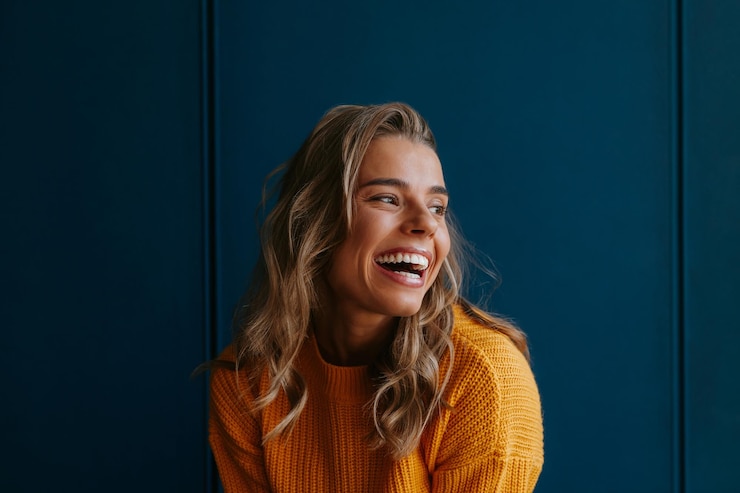 Foto gelukkige jonge blonde vrouw in een gele trui die wegkijkt terwijl ze op blauwe achtergrond staat