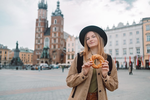 Gelukkige jonge blonde vrouwelijke toerist met stijlvolle kleding en een boho-hoed die bagel obwarzanek tradit eet