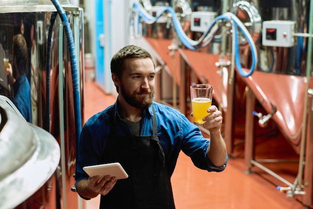 Gelukkige jonge brouwer met digitale tablet die na voorbereiding naar glas bier in zijn hand kijkt terwijl hij in het gangpad staat tegen enorme stalen tanks