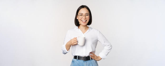 Gelukkige jonge, energieke aziatische vrouw die lacht en een kopje koffie drinkt, staat zelfverzekerd tegen een witte achtergrond