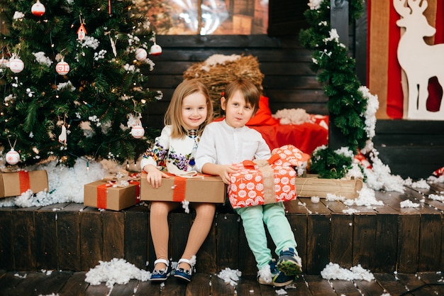 Gelukkige jonge geitjes die giftdozen in studio met cristmasboom en nieuwe jaardecoratie openen.