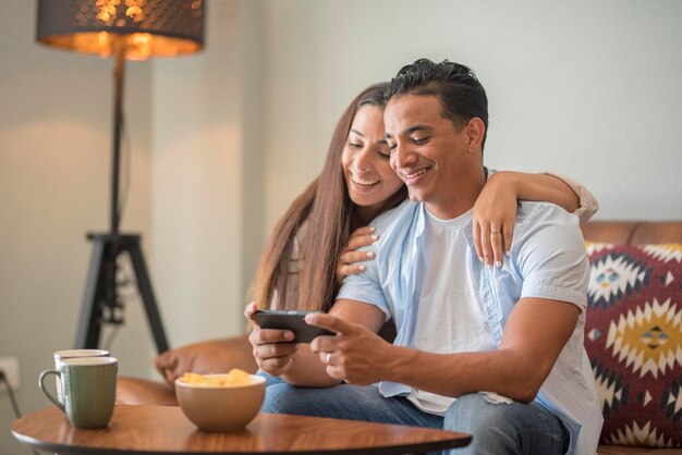 Gelukkige jonge gelukkige paar met behulp van smartphone sociale media-apps thuis lachende man en vrouw millennial gebruikers klanten praten hechting kijken naar grappige video kijken naar mobiele telefoon ontspannen op de bank