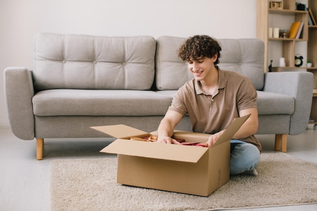 Gelukkige jonge glimlachende gekrulde man die doos opent met bestelde goederen geschenken cadeaus thuis op de bank Online shopper mannelijke klant die online winkelpakket opent