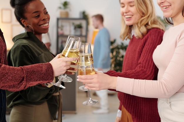 Gelukkige jonge interculturele vrouwen in nette vrijetijdskleding die klinken met champagneglazen terwijl ze plezier hebben op een thuisfeestje tegen hun vriendjes