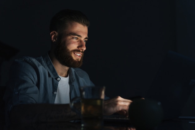 Gelukkige jonge man aan het werk op een laptop terwijl hij 's nachts binnenshuis aan tafel zit