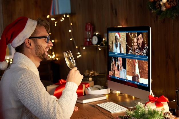 Gelukkige jonge man die een kerstmanhoed draagt, champagne drinkt, met vrienden praat via virtuele videogesprekken, bijeenkomt, nieuwjaarsfeest viert op vakantie, online conferentie, chat samen op de computer thuis.