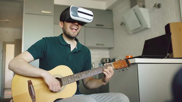 Gelukkige jonge man die in de keuken zit en gitaar leert spelen met een vr 360-headset