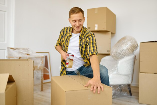 Gelukkige jonge man die kartonnen doos inpakt om te verhuizen naar een nieuw huis appartement dat in de woonkamer staat