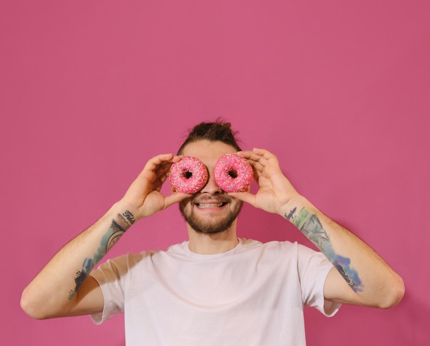 Gelukkige jonge man die lacht terwijl hij twee roze donuts voor zijn ogen houdt en plezier heeft