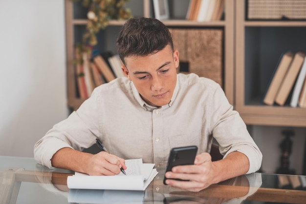 Gelukkige jonge man die zijn smartphone thuis gebruikt om op het net te surfen en huiswerk te maken met plezier alleen binnenshuis. Mannelijke tiener met telefoon geniet van vrije tijd studeren en werken.